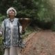 Petra Richarz von PERI Shamanic Spirit steht im Wald, hat die Klangschale gerade angeschlagen und lauscht dem Klang nach, während sie verbunden nach oben blickt; im Hintergrund führt ein Waldweg weiter in die Tiefe.