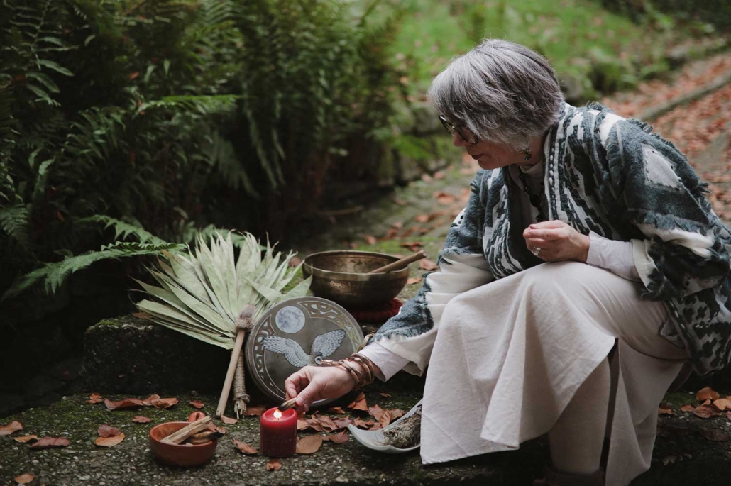 Petra Richarz von PERI Shamanic Spirit hockt im Wald und entzündet ein Stück Palo Santo an einer roten Kerze; im Hintergrund liegen Palmwedel, Trommel und Klangschale.